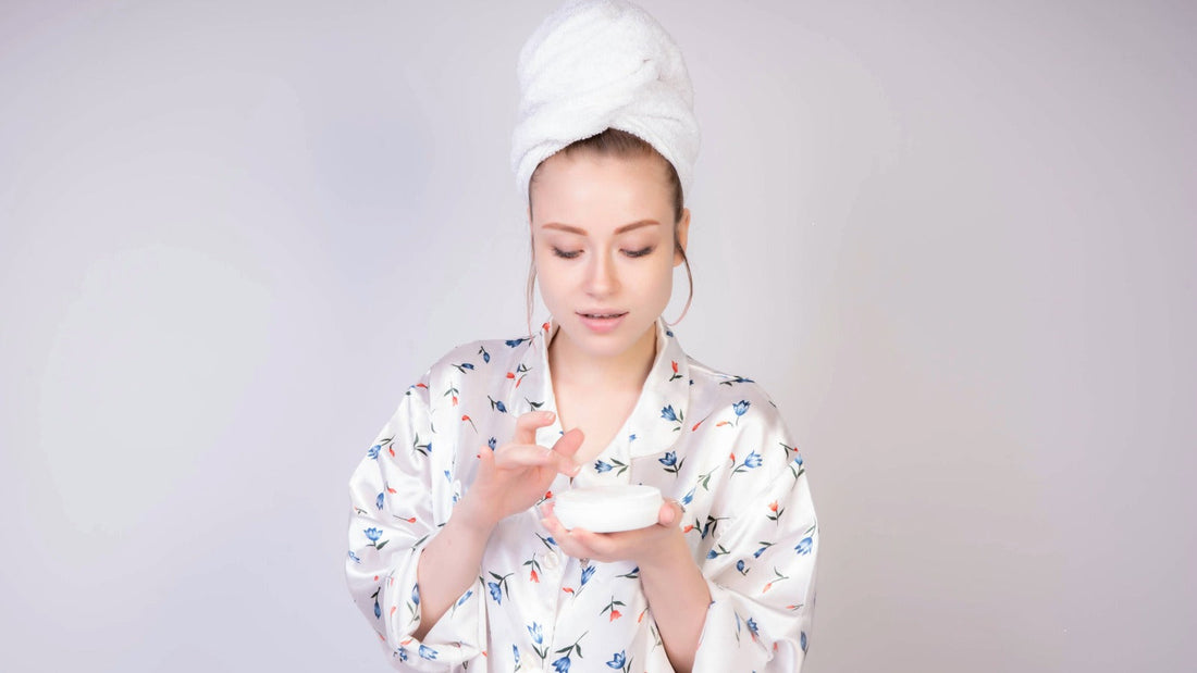 A woman in a towel wrap and floral robe applying skincare cream, representing a natural glow