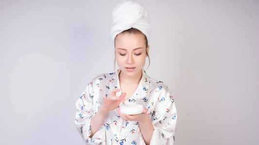 A woman in a towel wrap and floral robe applying skincare cream, representing a natural glow