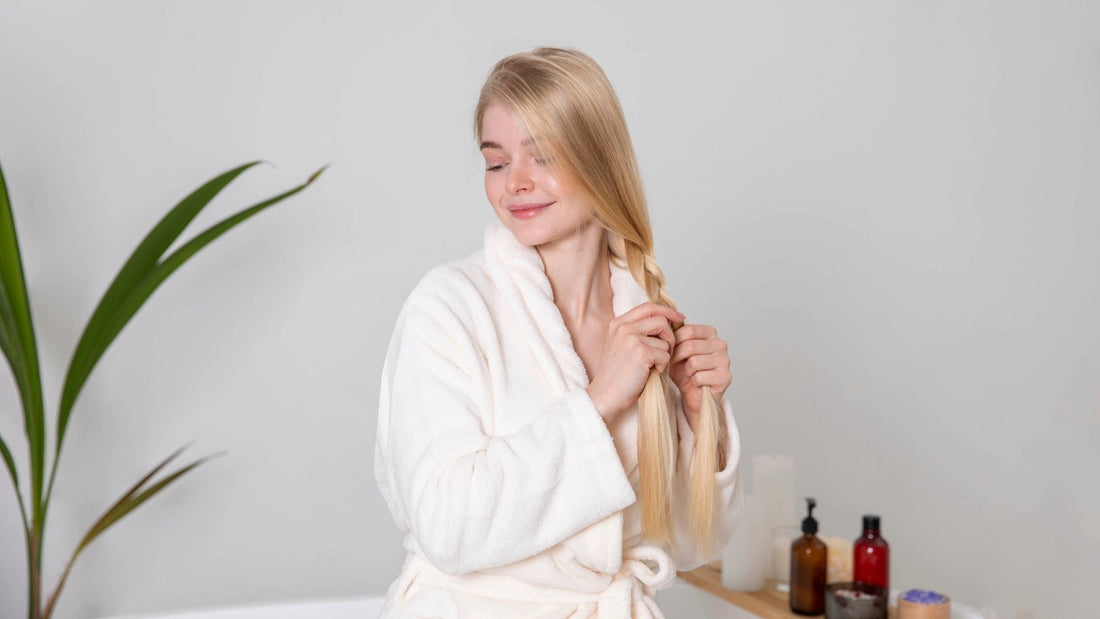 A woman in a white robe gently braiding her long blonde hair as part of her hair care routine.