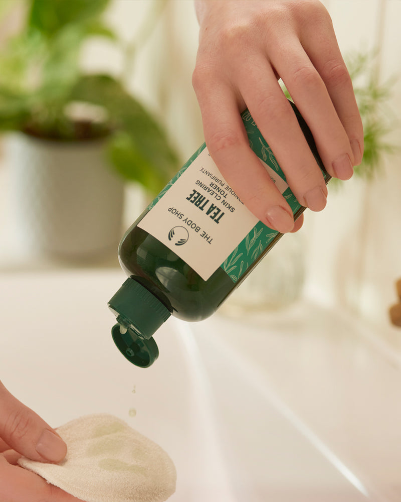Hand holding a green bottle with a label, likely containing a skincare product, with a blurred indoor background.